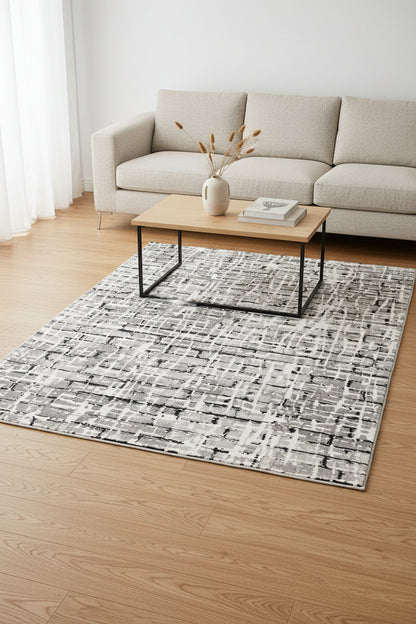 An abstract area rug featuring a modern, distressed grid pattern in shades of gray, black, and off-white.