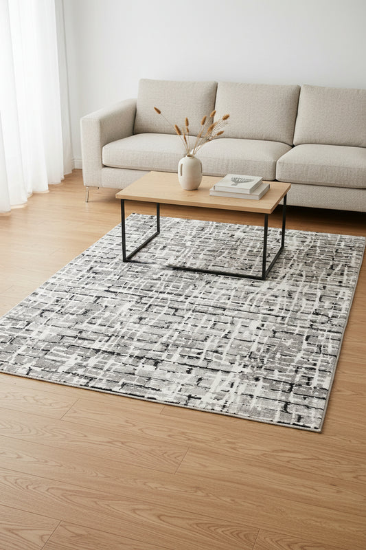 An abstract area rug featuring a modern, distressed grid pattern in shades of gray, black, and off-white.