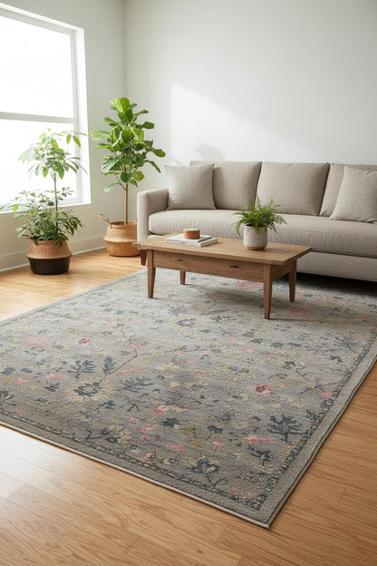 A rectangular vintage-style area rug with a distressed gray base and an intricate floral pattern in muted pink, beige, and dark blue-gray tones.